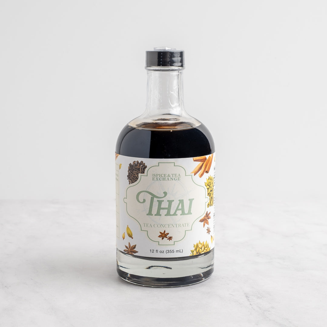 A bottle of Spice Tea Exchange Thai Leaf Concentrate, featuring a clear glass bottle with dark liquid inside and a white label decorated with tea and spice illustrations, set on a light marble surface against a white background.